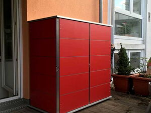 . The image shows a bright red storage unit or cabinet situated on a wooden deck. It is placed against the wall of a building, next to a door. There are some potted plants nearby. The storage unit appears to be outdoors, likely used for organizing garden tools or other items.