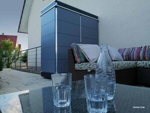 . Ein modernes Gartenmöbel-Set mit Gläsern auf einem Glastisch und einem blauen Aufbewahrungsschrank im Hintergrund.