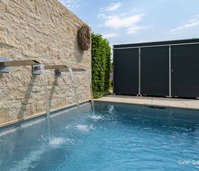 Ein moderner Pool mit Wasserfällen an einer Steinmauer, im Hintergrund steht ein großer Schuppen.
