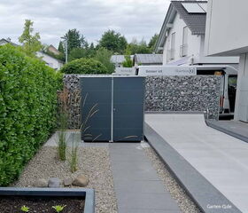 Moderner Gartenbereich mit Kies, Hecke und grauem Schrank, daneben ein Weg und Steinkorbzaun.