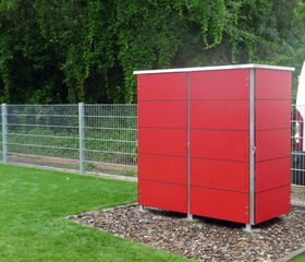. The image shows a red outdoor structure that resembles a storage cabinet or locker, with a metal frame. It is standing on a base of gravel, positioned on a grassy area next to a metal fence. There's some greenery in the background, and part of a white vehicle is visible on the right.