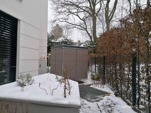 Ein verschneiter Garten mit Zaun, kahlen Bäumen, einem Schuppen und einem modernen Gebäude.