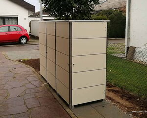 Ein moderner Garten- oder Geräteschrank steht in einem Vorgarten neben einem roten Auto auf der Straße.