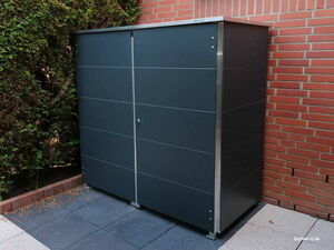 . Ein moderner, dunkelgrauer Gartenschrank steht auf einer Terrasse neben einer Ziegelwand und Hecken.
