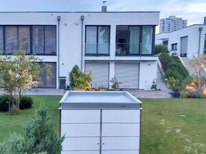 . The image shows a modern, white storage unit or garden shed situated on a grassy area in front of a contemporary house. The house has large windows, some with balconies, and a patio area. There are plants and trees around, with the lawn leading up to the house. The setting appears to be a modern residential area.