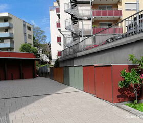 Mehrere moderne Gebäude mit Balkonen, daneben bunte Müllcontainerboxen und Pflanzen in einem gepflasterten Innenhof.