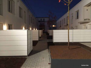 . Moderne Wohnanlage bei Nacht mit Zäunen, gepflastertem Weg, beleuchteten Gebäuden und Bäumen.