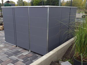 . The image shows a grey outdoor structure, possibly a storage unit or garden shed, on a paved area. There are plants nearby and a few people dressed in what appears to be work or gardening attire. The scene seems to involve some landscaping or garden work.