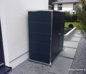 . Metallschrank neben einem Haus auf einer Kiesfläche, hinter einem Weg und Gartenbeet.