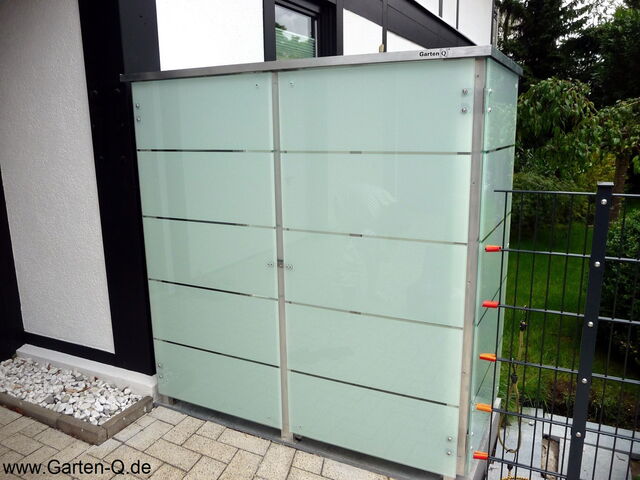 . The image shows a sleek, modern storage unit with a frosted glass exterior, reinforced by metal frames. It's placed outdoors on a tiled patio next to a building, with some white stones neatly arranged near the base. There are some orange hooks or fixtures attached to the side, and a green lawn is visible in the background. The unit is labeled with "Garten-Q" on top.