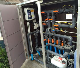 The image shows an equipment cabinet or control panel with multiple pipes and valves, likely part of a plumbing, water filtration, or pool system. It includes various PVC pipes, orange-handled valves, and electrical components mounted on the interior wall. There is also a container with a liquid, possibly for chemical treatment or cleaning.