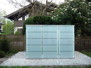 . The image shows a modern, frosted glass structure situated in a garden setting. Behind it, there's a wooden house, some trees, and shrubs. The ground is paved with stone tiles.