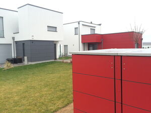 . The image shows a modern housing complex with minimalist architectural design. The buildings feature a combination of clean lines and flat roofs. The color scheme includes white, red, and dark gray panels. There's well-maintained grass and landscaping in the foreground.