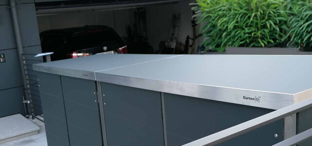 Garage mit Auto, daneben ein Garten-Q Schrank mit SmartDach aus HPL-Trespa-Platte, umgeben von Grünpflanzen.