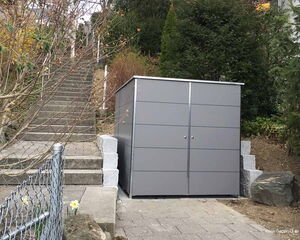 . Graues Gartenhaus aus Metall neben einer Treppe, umgeben von Bäumen und Pflanzen.