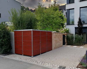Ein Gartenschuppen aus Holz neben einem modernen Gebäude, umgeben von Bäumen und Sträuchern.