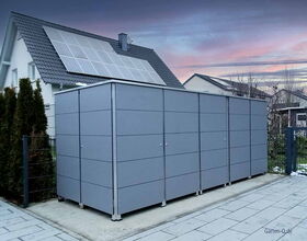 Graues Gartenhaus neben einem Haus mit Solarpanelen auf dem Dach und umgeben von Bäumen.