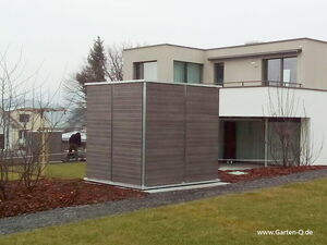. The image features a modern house with a rectangular structure in the foreground that appears to be a garden shed or storage unit. The structure has a minimalist design with wooden panels. The house is two-story, with large windows and a flat roof. The surrounding area includes a pathway, grass, and some barren trees or bushes, indicating a possibly early-spring or late-autumn setting.