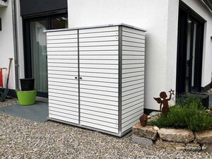 . Ein weißer Gartenschrank steht auf einer Kiesfläche neben einem Haus, mit Pflanzen und einer Engel-Deko daneben.