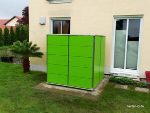Ein grüner Gartenschrank steht neben einem Haus, umgeben von Gras und einer Palme.