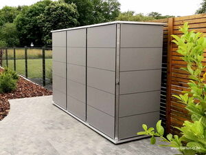 . The image shows a sleek, modern garden storage shed with a metal finish. It's placed on a paved area with a wooden fence on one side and trees in the background. A fenced field is visible beside it, along with some landscaping and plants nearby.