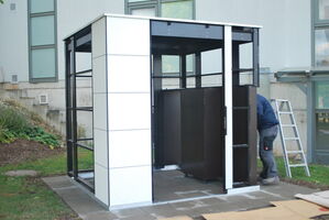 The image shows a partially constructed metal and panel structure resembling a small outdoor building or shelter. It appears to have a framework made of metal, with panels covering some areas. There is a person working on the structure, and a ladder nearby. The setting is outdoors, with grass and a building in the background.