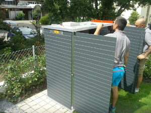 . The image shows two people assembling or installing a metal structure, possibly a storage shed or enclosure, outdoors. The body of the structure is made of horizontal slats and stands on a paved surface. There's a level tool placed on top, and the surrounding environment includes grass, a fence, and some trees.