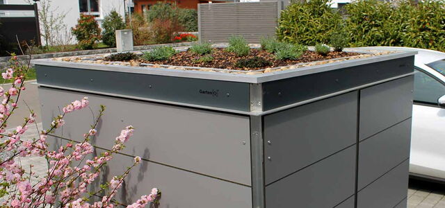 Ein grauer Gartenschrank mit bepflanztem Dach, umgeben von blühenden Büschen, in einem modernen Wohngebiet.