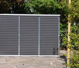 . The image shows a large solar panel placed outdoors. It is standing upright on a paved surface, surrounded by greenery and plants. There is a wooden post and shrubs nearby.