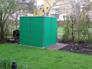 . The image shows a green garden shed or storage unit situated in a garden area. There are trees and shrubs around it, and a grassy lawn in the foreground. To the left, there is a bicycle partially visible next to the shed. Additionally, there's a path made of bricks or tiles leading to or around the shed.