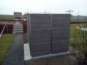 . This image shows an outdoor storage shed with a wooden-like finish. It's placed on a paved area surrounded by grass. There are a few other structures and features visible in the background, such as steps leading up a small hill and some kind of a frame or board further behind. The setting appears to be a garden or backyard area.