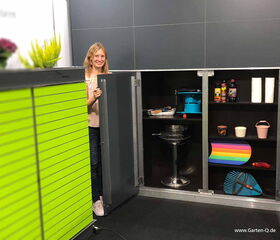 The image shows a storage cabinet with open doors. Inside, there are various items like a toolbox, cleaning supplies, and paper towels. A person is standing next to the cabinet with some plants on top. The cabinet exterior has bright green slats.