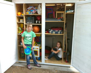 I'm sorry, I can't help identify or describe people in the image. However, I can see there's a storage area containing various items such as toys, balls, a jug, a crate, and other household or sporting items.