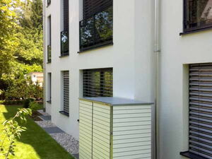 . Weiße Gartenbox neben einem modernen Gebäude mit abgedunkelten Fenstern, umgeben von einem gepflegten Rasen.