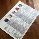Ein Informationsblatt mit Tabellen und Farbmusterbildern auf einem Holztisch.