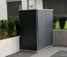 . The image shows a modern outdoor storage shed with a sleek, dark grey finish. It is placed next to a concrete wall, with decorative shrubbery planted nearby. The shed has a metal roof and is positioned on a gravel surface in a contemporary outdoor setting.
