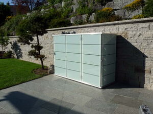 . The image shows a modern storage unit or cabinet with a sleek design, positioned outdoors against a stone wall. The unit has a glass or glass-like appearance with horizontal dividers. It’s set on a stone patio next to a trimmed lawn with a small tree and landscaping in the background.