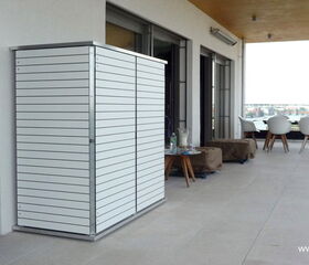 . The image features an outdoor balcony or terrace area. There is a modern white storage cabinet with horizontal lines on the left side. The area includes seating with a few chairs and tables. The setting appears to be minimalistic and contemporary, with large windows and a view of the outdoors.