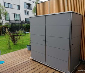 . Ein grauer Gartenschrank auf einer Terrasse, daneben ein Garten mit Trampolin und mehrstöckigem Gebäude im Hintergrund.