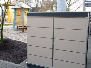. Ein grauer Schrank im Freien, davor ein Spielplatz mit Holzspielhaus und Sandboden.