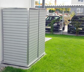 . The image shows an outdoor air conditioning unit enclosure made of horizontal slats positioned against the exterior wall of a building. It is surrounded by a grassy area. In the background, there are two parked cars behind a wire fence.