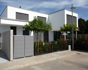 . The image shows a modern, two-story building with a minimalist design. It features clean lines and a predominantly white facade. The windows have horizontal slats, and there are two small trees with pruned canopies in front of the building. A streetlight is also visible, along with a fence and some landscaping that includes shrubs. The sidewalk and a paved driveway or path are in the foreground.