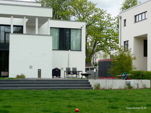. The image shows a modern home with a white exterior and large windows. In front of the house, there's a patio with a table and chairs, a grill, and an umbrella. A grassy lawn with steps leads up to the patio. There are trees and bushes surrounding the area, along with a lawnmower and a toy in the garden.
