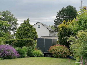 . Ein gepflegter Garten mit verschiedenen Büschen, Bäumen und einem modernen Schuppen im Hintergrund.