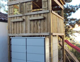 . The image shows a wooden structure, resembling a small playhouse or lookout tower, situated above a cabinet or storage unit. The playhouse has an open design with railings and a roof, and there's a ladder or slide leading to or from it. It's positioned outdoors, surrounded by trees and a house.