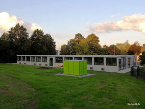 . Ein modernes Gebäude mit vielen Fenstern, umgeben von grüner Wiese und Bäumen, hat ein großes, grünes Quadrat davor.