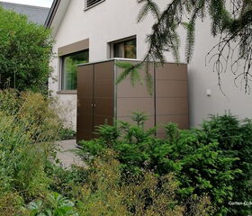 . Ein brauner Gartenschrank steht neben einem Haus in einem üppig begrünten Garten.