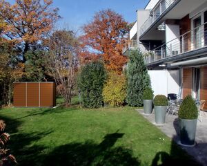 The image shows a modern residential building with a garden area. In the garden, there is a patch of green grass, shrubs, and trees with autumn foliage. To the left, there is a wooden structure that looks like a shed or storage unit. On the patio, there are several large potted plants and some outdoor furniture.