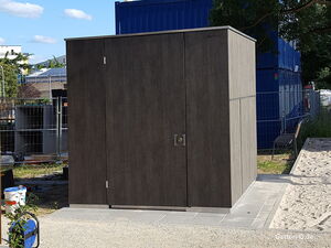 . Ein modernes Gartenhaus aus dunklem Holz mit Metallzaun und blauen Containern im Hintergrund.