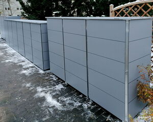 Graue, moderne Mülltonnenboxen auf gepflastertem Weg, umgeben von Pflanzen und leichter Schneedecke.